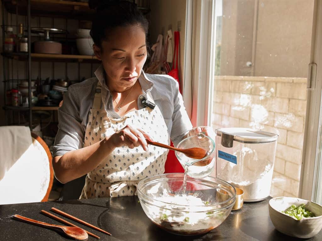 A woman wearing an apron pours water into a mixing bowl of flour while preparing food in a sunlit kitchen, with cooking utensils and ingredients on the table.